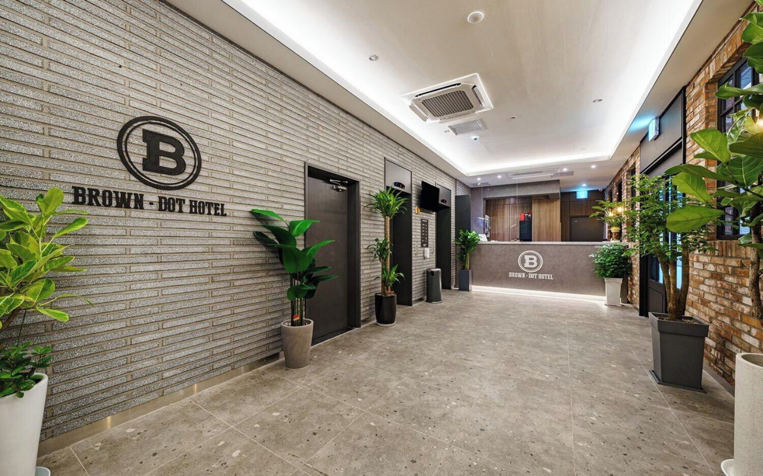 A hotel reception area with a modern design, featuring a brick wall, a reception desk, and potted plants.