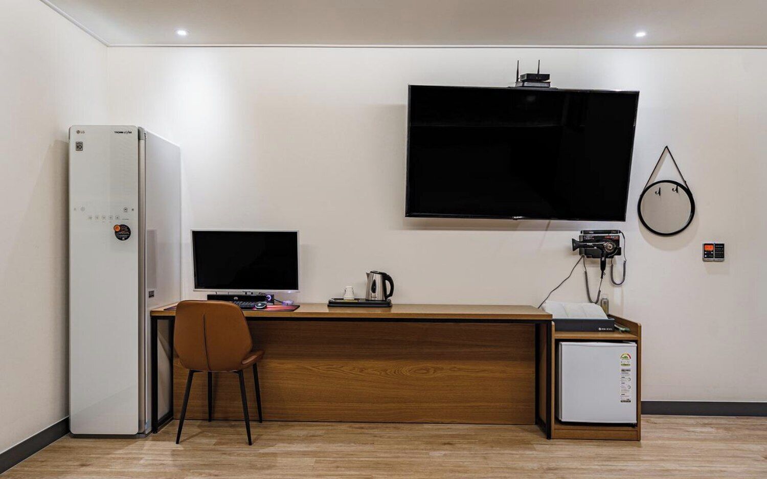 A hotel room with a wooden desk, a television mounted on the wall, a refrigerator, and a small cabinet.
