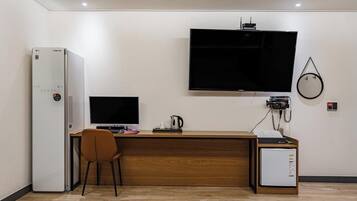 A hotel room with a wooden desk, a television mounted on the wall, a refrigerator, and a small cabinet.