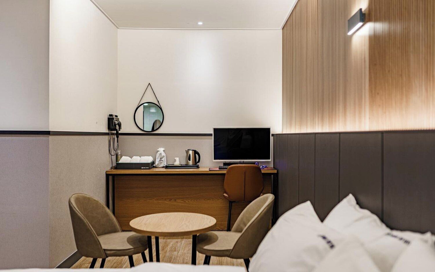 Habitación de hotel con cama, dos sillas, una mesita redonda, un escritorio con computadora, un espejo y una lámpara de pared.