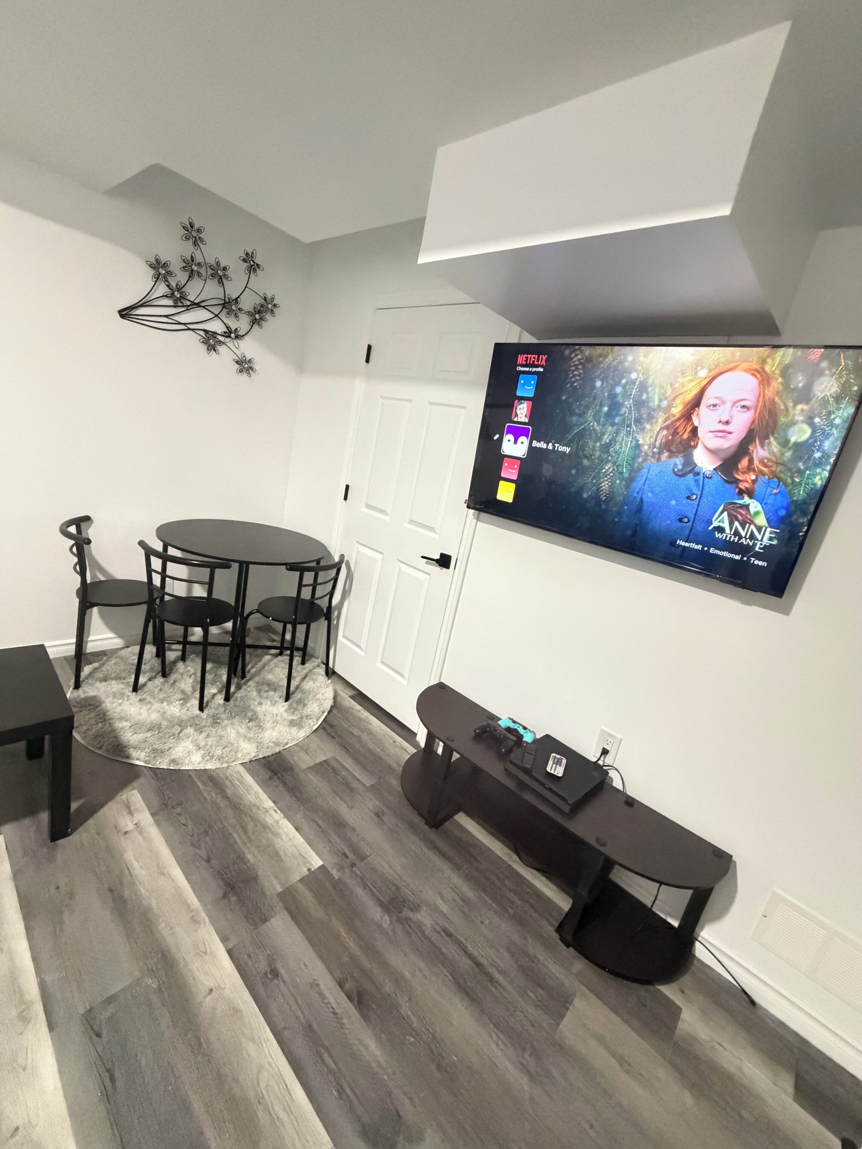 Un salon moderne avec un téléviseur à écran plat fixé au mur, une table à manger ronde avec des chaises, un meuble de rangement noir et un tapis moelleux sur le plancher en bois.
