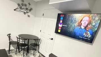Een moderne woonkamer met een flatscreen televisie aan de muur, een ronde eettafel met stoelen, een zwarte entertainmentkast en een zacht vloerkleed op de houten vloer.