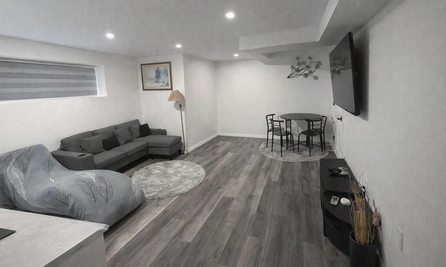 A modern living room with a grey sectional sofa, a round dining table with chairs, a floor lamp, and a flat-screen TV mounted on the wall.