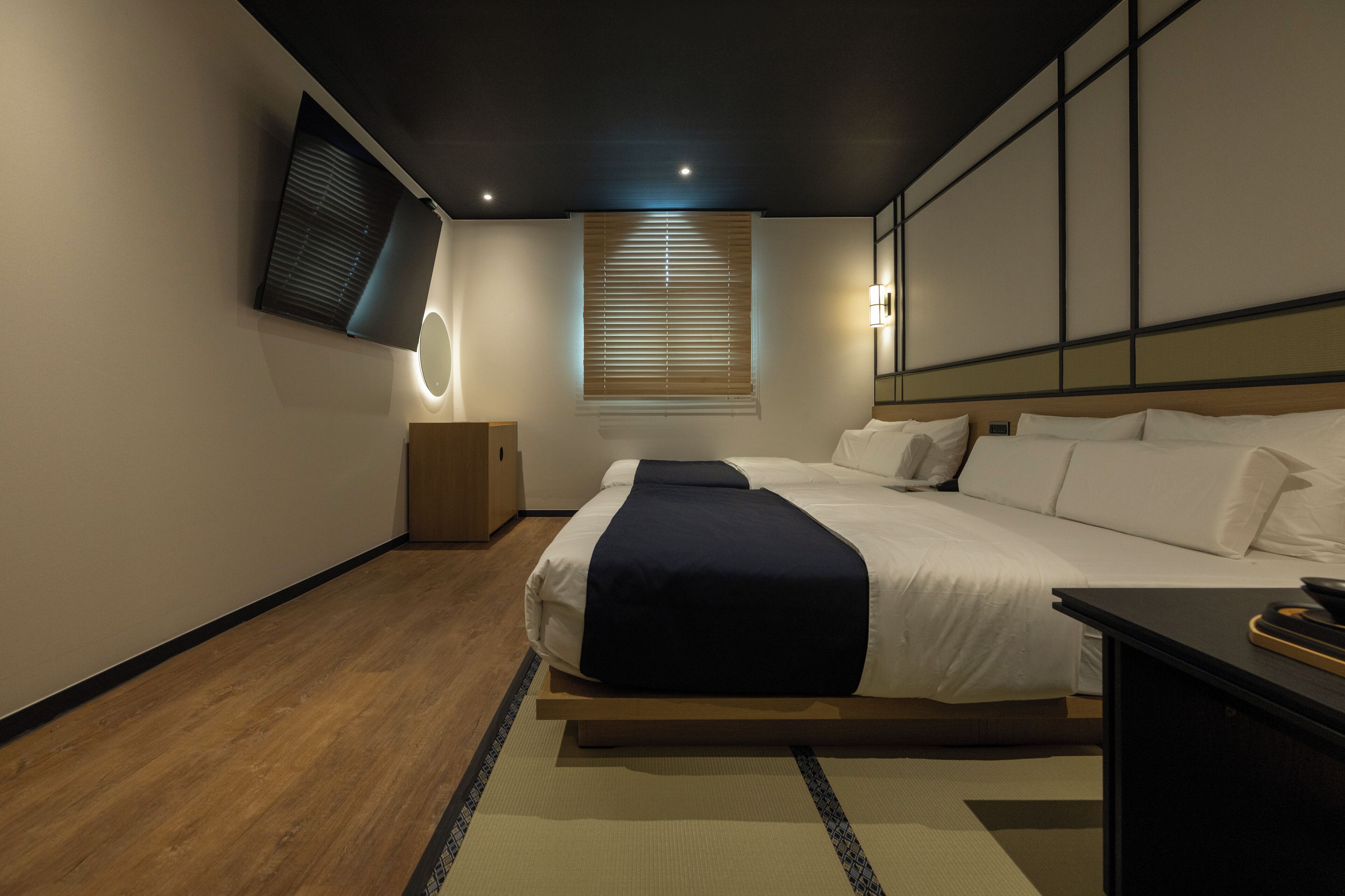 Una moderna camera d'albergo con un letto grande, una TV a schermo piatto montata a parete, un comodino e una finestra con persiane.
