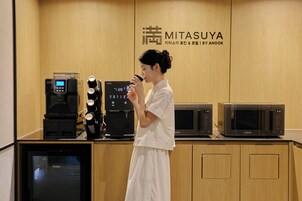 Una persona con vestido blanco de pie frente a una exhibición de electrodomésticos de cocina, incluyendo una cafetera, un microondas y una cocedora de arroz.