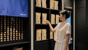 A woman in a white dress examining packaged items on shelves in a store with a blue cloth and wooden balls in the background. - Mitasuya Ryokan Dongducheon Soyosan (Dongducheon)