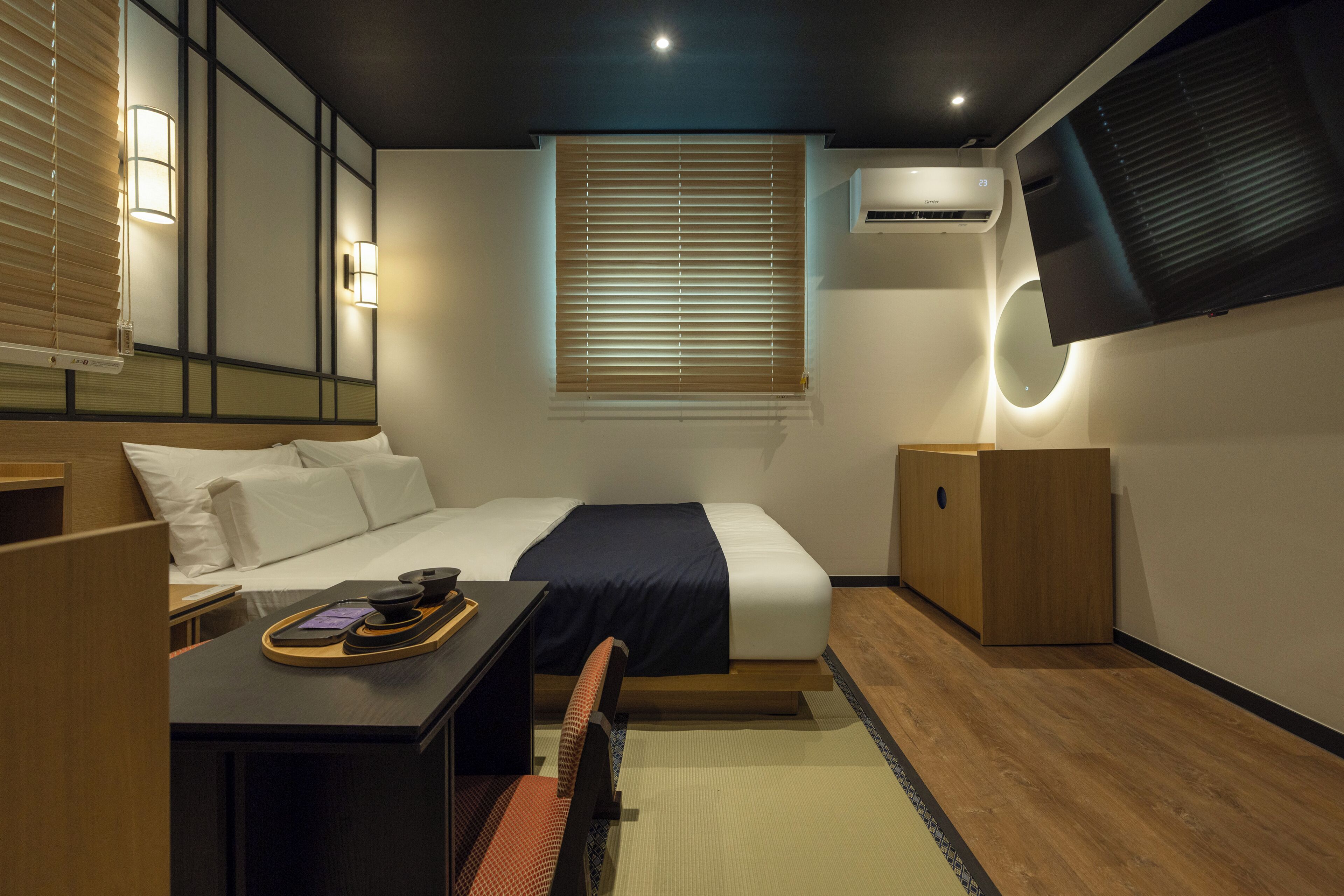 A hotel room with a bed, bedside table, chair, desk, and a TV mounted on the wall.