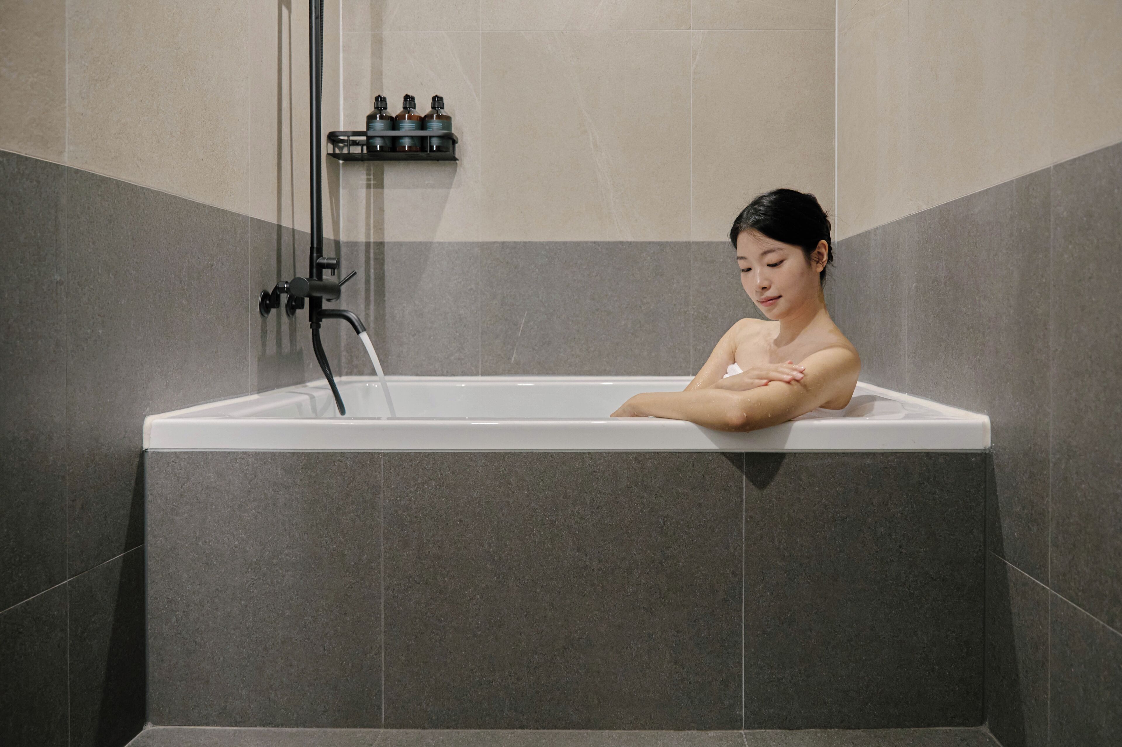 A person relaxing in a bathtub with a water faucet and a shelf with bottles.