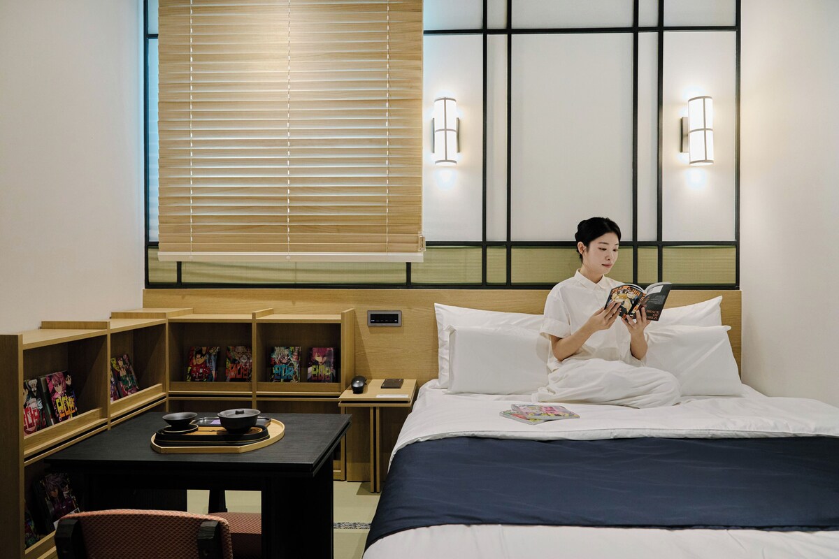Habitación de hotel con cama, mesita de noche, lámpara de lectura y una pared con ventana y persianas.