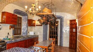 A kitchen with patterned tiles, wooden cabinets, a hanging pot rack, and a dining area with a patterned tablecloth.