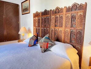 Una cama con una colcha estampada, dos almohadones decorativos, un cabecero de madera con intrincados grabados, una mesita de noche con una lámpara y un armario de madera.