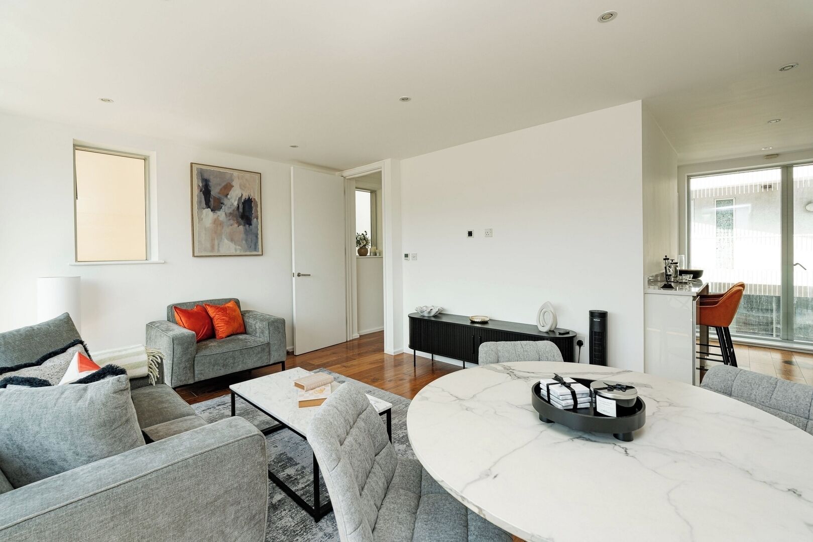 A modern living room with a grey sofa, a round marble table, and a kitchen area with a black cabinet.