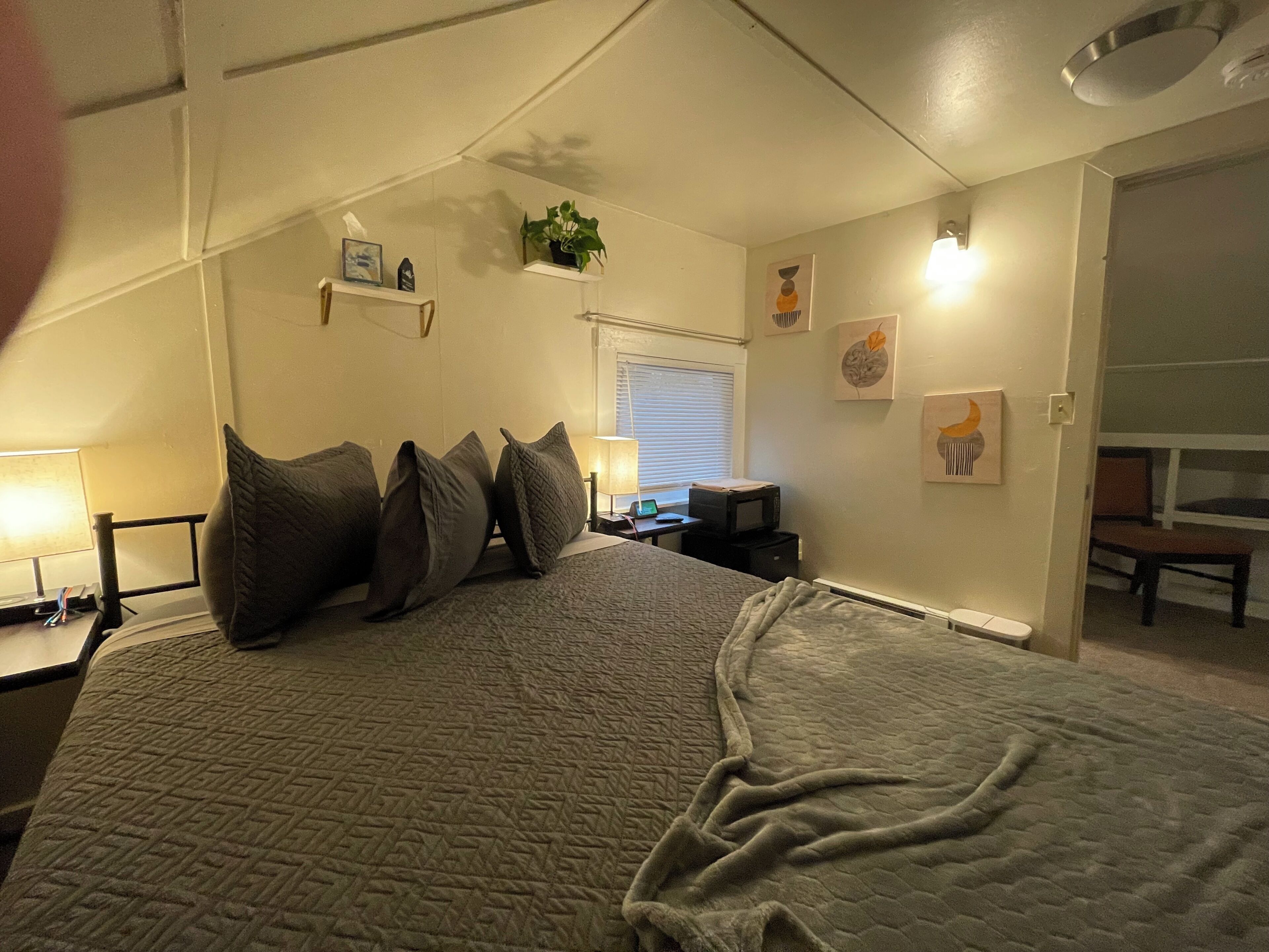 A bedroom with a bed, bedside tables, a desk with a computer, a window with blinds, and wall art.