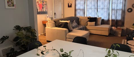 A living room with a sofa, armchairs, a coffee table, and a dining table. There is a door with a coat rack and a plant on the wall.