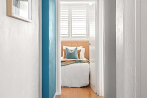 A narrow hallway leading to a bedroom with a bed, pillows, and a window with blinds. - Retro Coastal Retreat w Pool and Kings Beach Views (Caloundra Region)