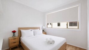 A modern hotel room with a large bed, wooden headboard, a nightstand with a lamp, and a window with blinds. - Spacious 3-Bed Townhouse Near Broadwater (Labrador)