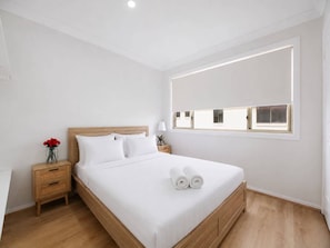 Ein modernes Hotelzimmer mit einem großen Bett, einem Holz-Kopfteil, einem Nachttisch mit Lampe und einem Fenster mit Jalousien.