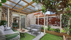 A covered patio area with a wooden pergola, a seating area with a table and chairs, and a view of a brick building in the background. - Spacious 3-Bed Townhouse Near Broadwater (Labrador)