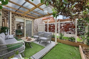 Ein überdachter Patio-Bereich mit einer Holz-Pergola, eine Sitzecke mit Tisch und Stühlen sowie ein Blick auf ein Backsteinhaus im Hintergrund.