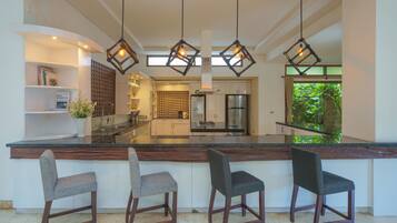 A modern kitchen with a central island, bar stools, pendant lighting, and a view of the greenery outside.