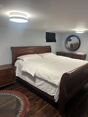 Ein Schlafzimmer mit einem hölzernen Bett, Nachttischen, einem runden Spiegel und einem kleinen Fenster mit Jalousien.