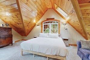 A bedroom with a sloped ceiling, a bed, a dresser, a window with a view, and a lamp.