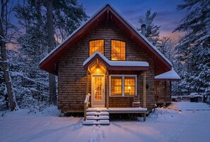 A wooden cabin with a snow-covered roof, a front porch, and a chimney.