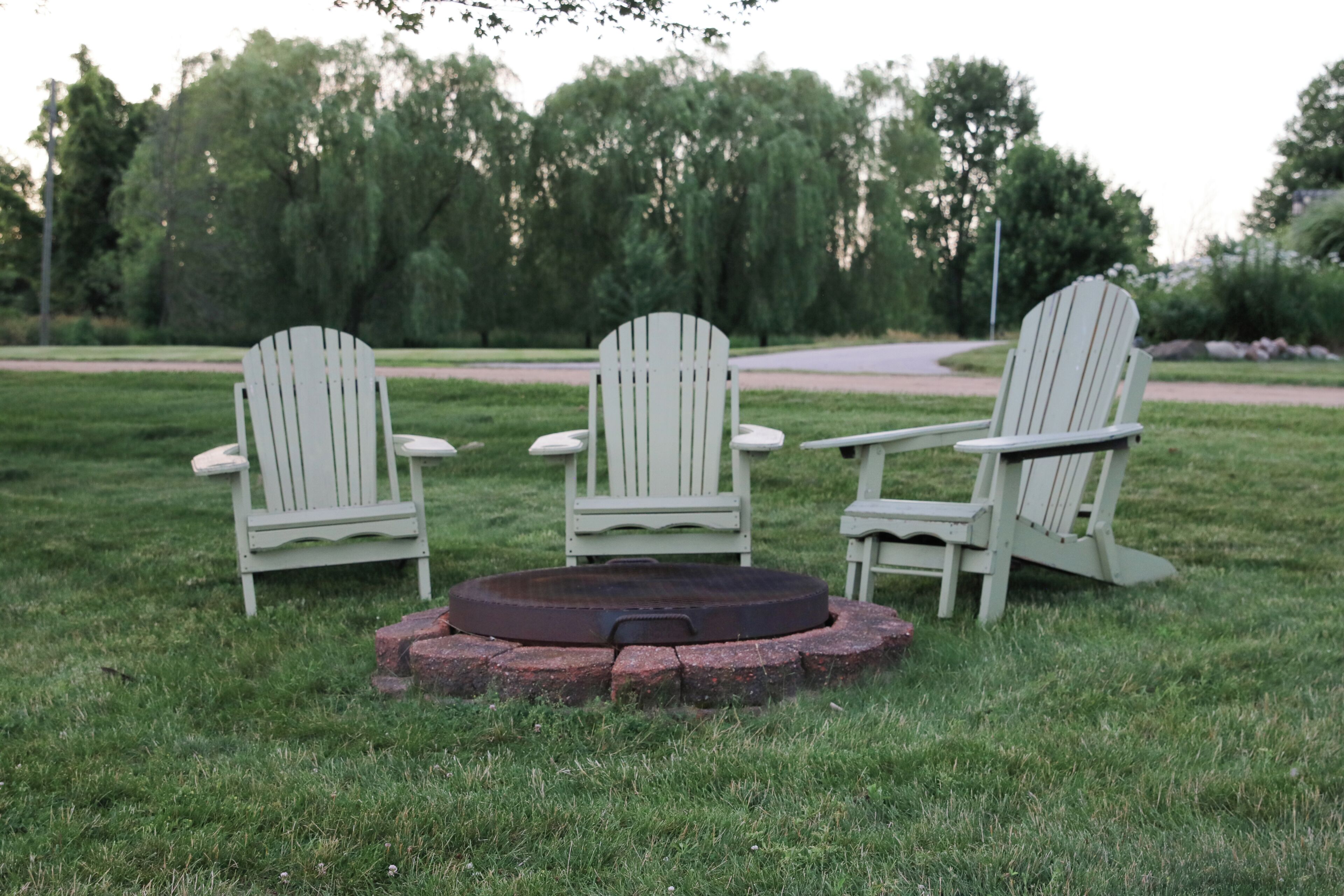 Three Adirondack chairs arranged around a circular fire pit on a grassy lawn with trees in the background.