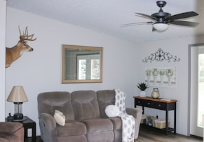 Un salon avec un canapé gris, un ventilateur de plafond, une décoration représentant une tête de cerf, un miroir et une œuvre d’art florale accrochée au mur.