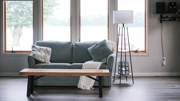 A room with a sofa, a floor lamp, and a table. There are three windows with blinds and a view of trees outside. - Enjoy a pleasant 3-bedroom Lake House in Scottville besides St Mary's Lake. (Scottville)