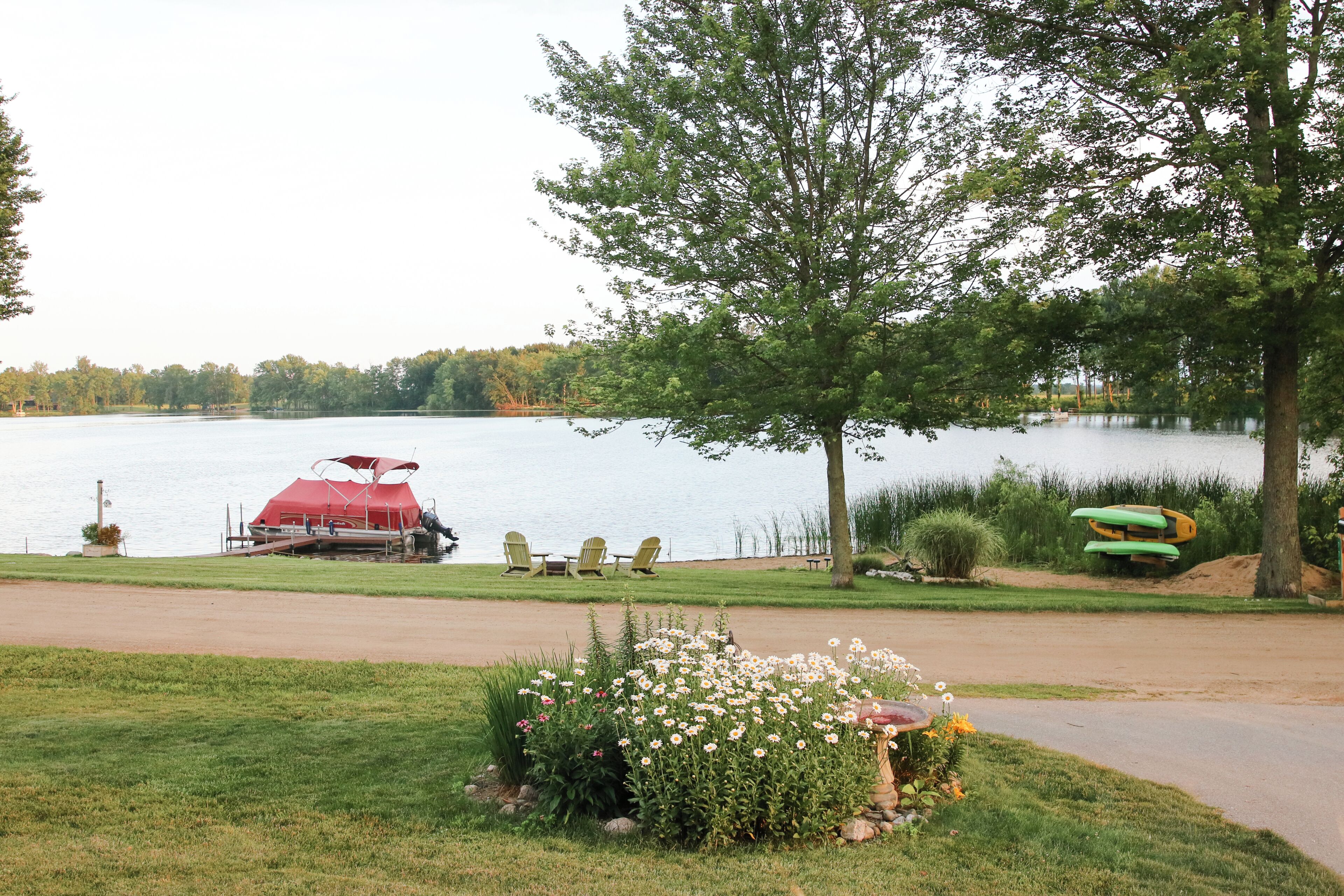 Un entorno junto al lago con un bote rojo amarrado, una hilera de sillas Adirondack y un pequeño jardín con flores.