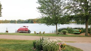 Un entorno junto al lago con un bote rojo amarrado, una hilera de sillas Adirondack y un pequeño jardín con flores.
