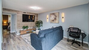 Uma sala de estar moderna com televisão de tela plana, um sofá azul, uma escrivaninha com cadeira e um grande quadro abstrato na parede.