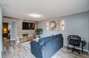 Ein modernes Wohnzimmer mit Fernseher, blauem Sofa, Schreibtisch mit Stuhl und einem großen abstrakten Gemälde an der Wand.