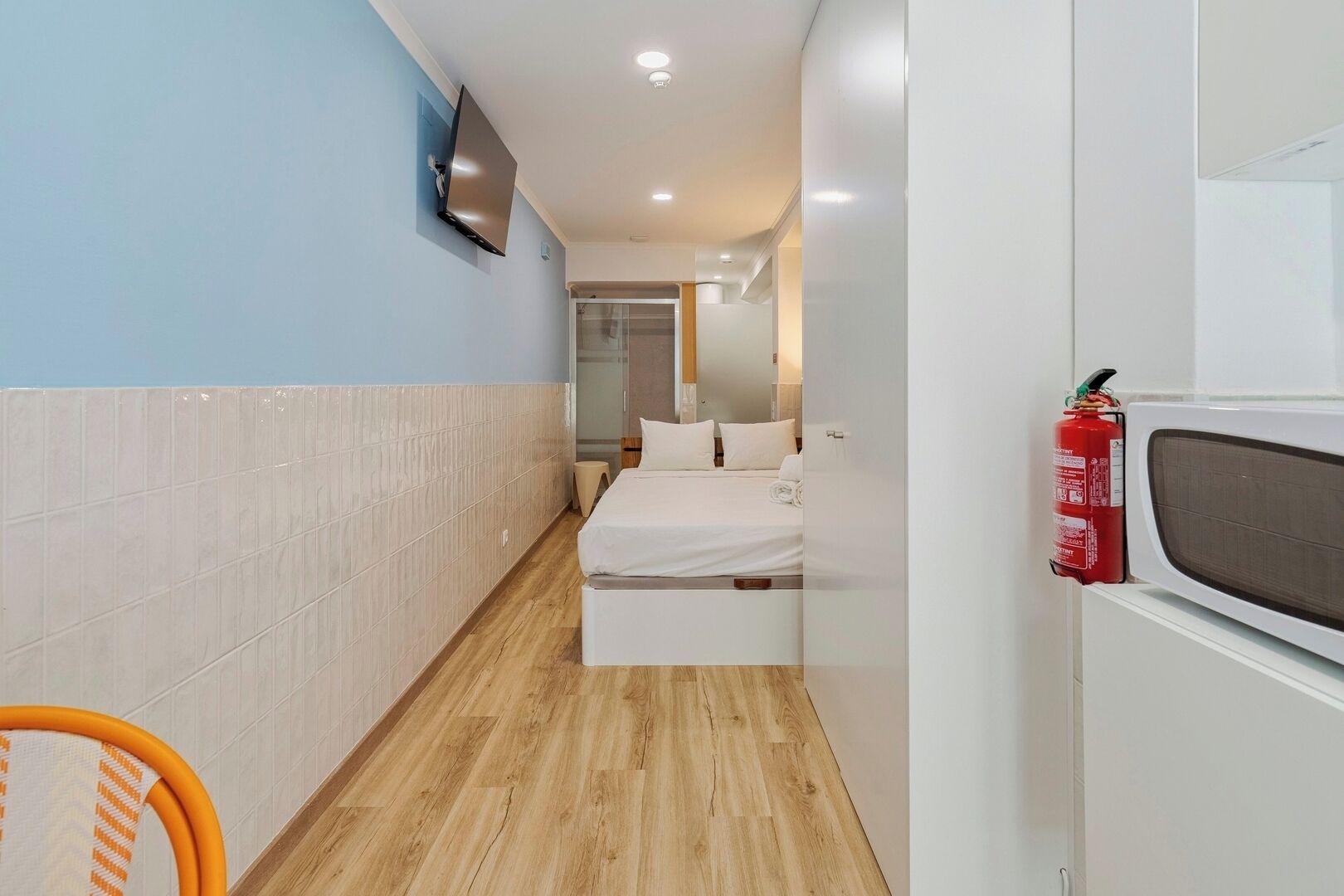 A compact hotel room with a bed, a television mounted on the wall, a fire extinguisher, and a microwave.