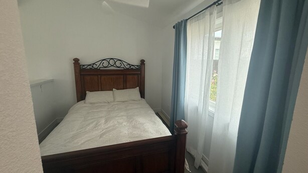 A single bed with a wooden headboard in a room with a ceiling fan, a window with blue curtains, and a view of greenery outside. - Minimalist 1 BR ADU in a pleasant Woodland neighborhood (Woodland)