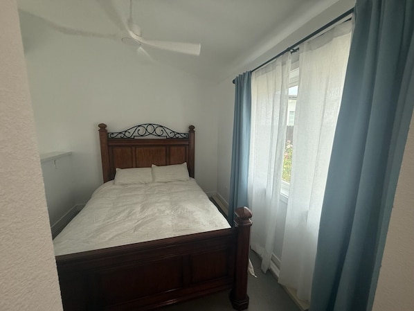 A single bed with a wooden headboard in a room with a ceiling fan, a window with blue curtains, and a view of greenery outside.