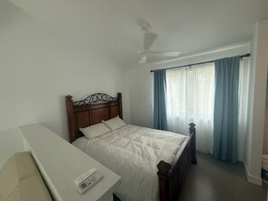 Quarto com uma cama de solteiro, mesa de cabeceira, ventilador de teto e janela com cortinas.