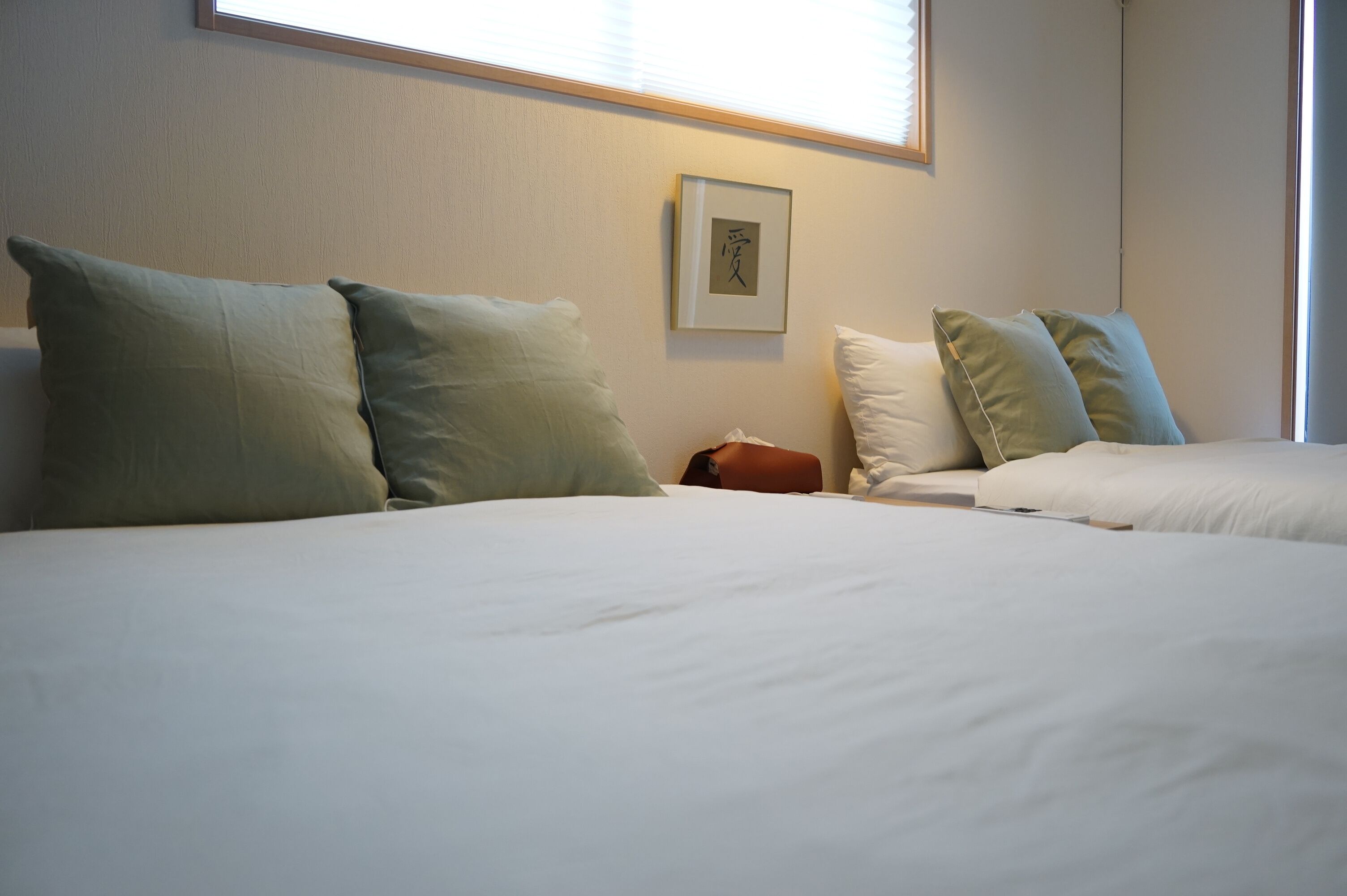 A hotel room with two beds, each with white bedding and green pillows, a framed picture on the wall, and a window with blinds.