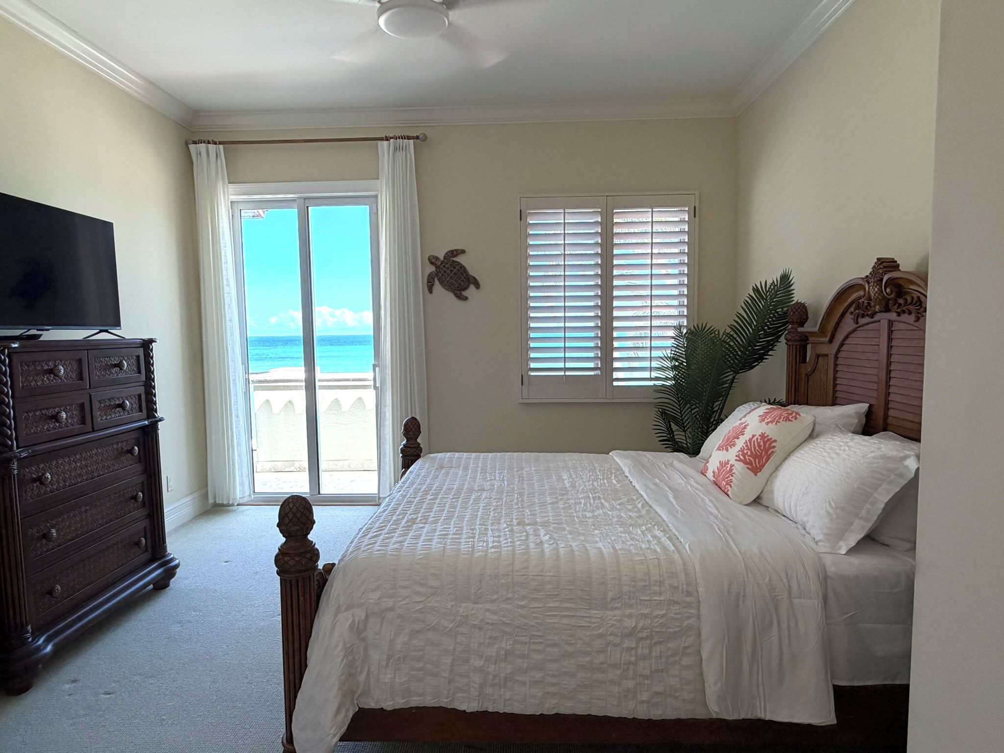 A bedroom with a bed, a dresser, a television, a window with shutters, and a view of the ocean.