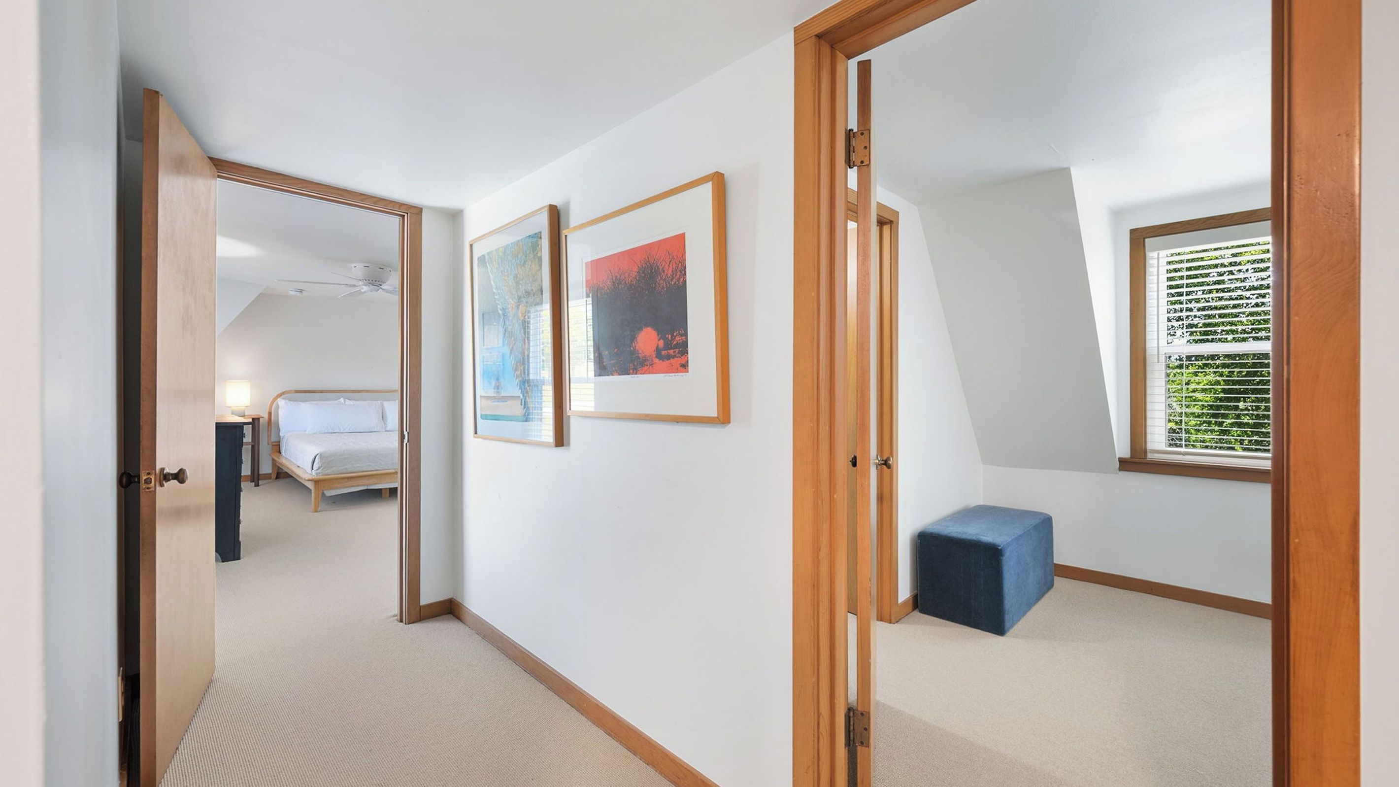 A hallway leading to a bedroom with a bed, framed artwork, and a window with blinds.