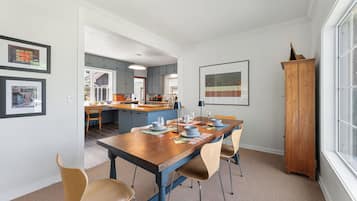 Uma sala de jantar com mesa de madeira posta para quatro, cozinha ao fundo e uma janela grande à direita.