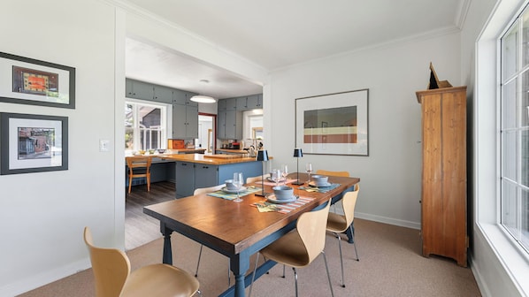 A dining area with a wooden table set for four, a kitchen in the background, and a large window on the right.