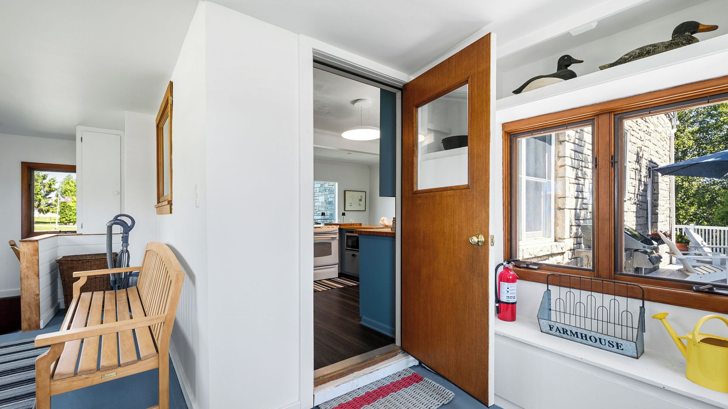 Quarto com um banco de madeira, porta que dá para uma cozinha e janela com vista para um edifício.