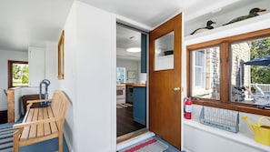 A room with a wooden bench, a door leading to a kitchen, and a window with a view of a building.