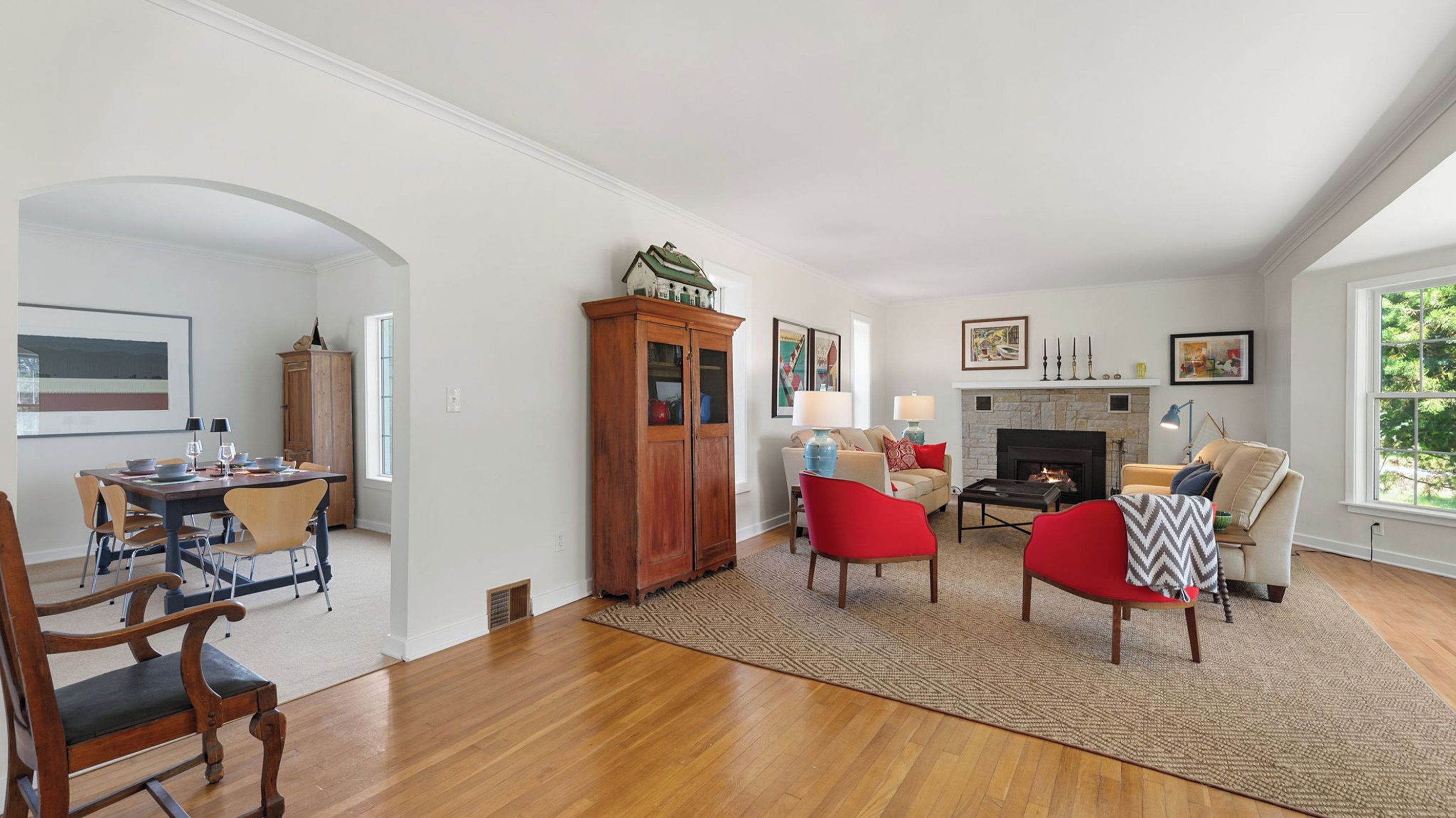 A spacious living room with a fireplace, red chairs, a dining area with a wooden table, and a view of greenery outside.
