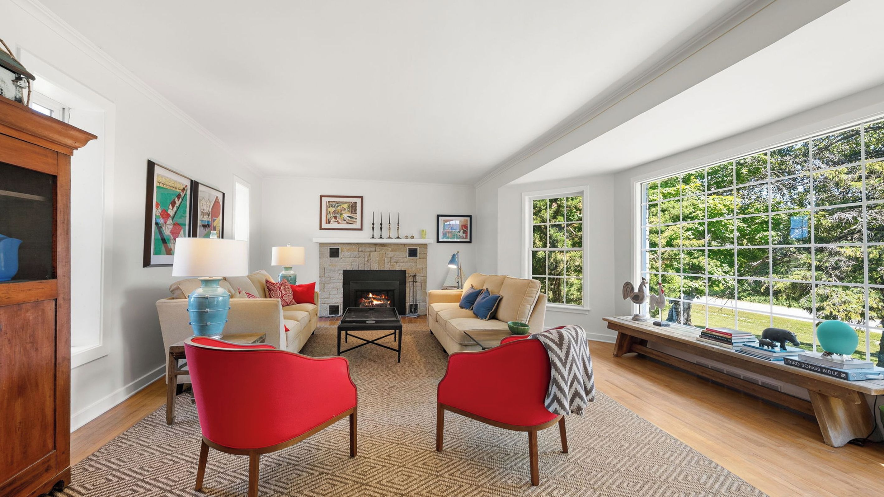 A living room with a fireplace, two red chairs, a sofa, and a window with a view of greenery.