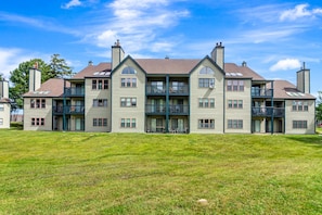 A multi-story residential building with a flat roof, balconies, and a grassy front yard.
