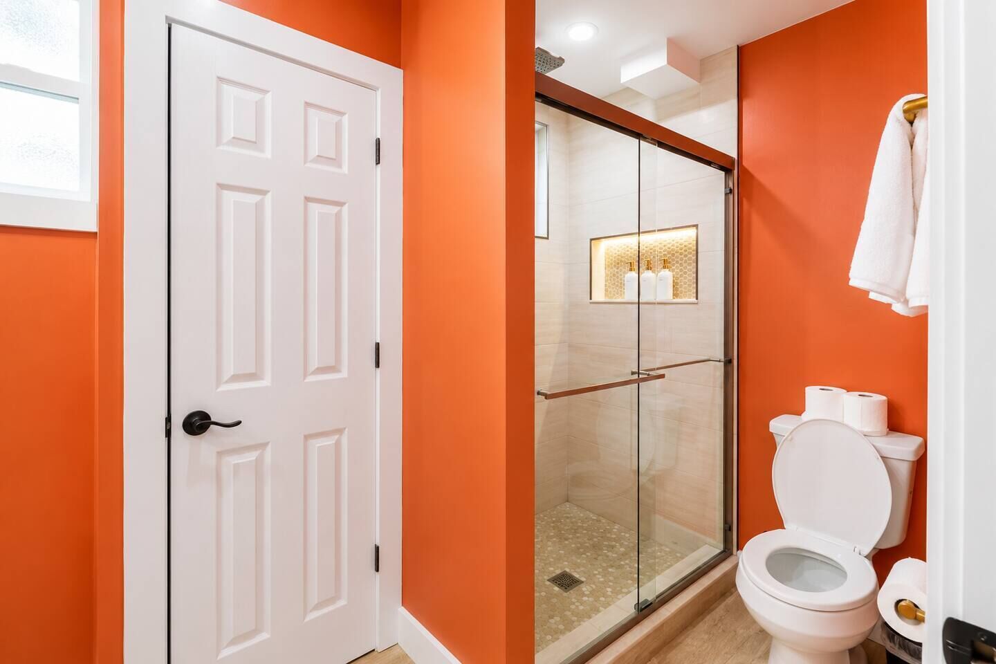 A bathroom with an orange wall, a white door, a glass-enclosed shower, and a toilet.