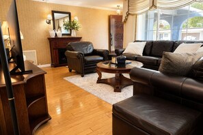 A living room with a leather sofa, a coffee table, a fireplace, and a large window with curtains. - Memories Beach Villa! Near Gulf Beaches + BBQ (Largo)